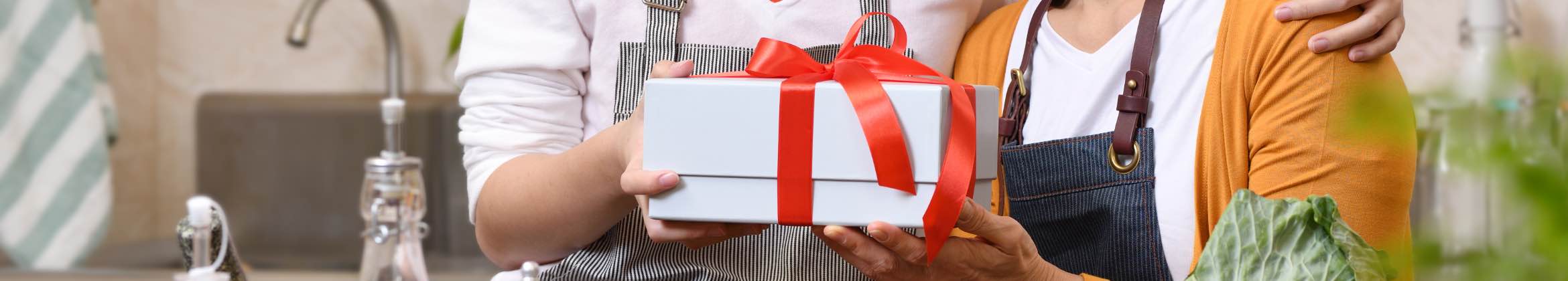 A person in an apron gives a wrapped gift to another person in an apron. In the foreground is a head of lettuce.