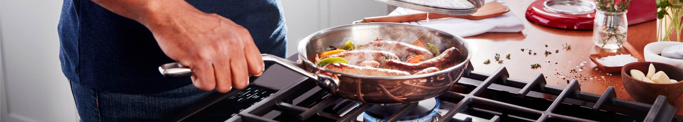 Someone cooks sausages in a pan on a KitchenAid gas cooktop. They hold the lid over over the pan. On the counter is a wooden spatula on a white dish cloth and some green herbs. 