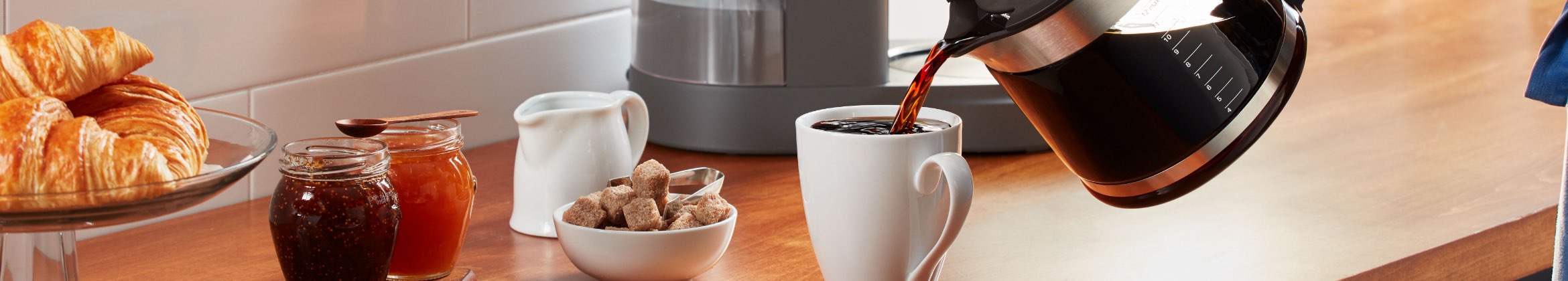 Coffee is being poured into a white mug on a counter. Also on the counter is a bowl of sugar cubes, two jars of honey, a drip coffee machine and a container of milk.