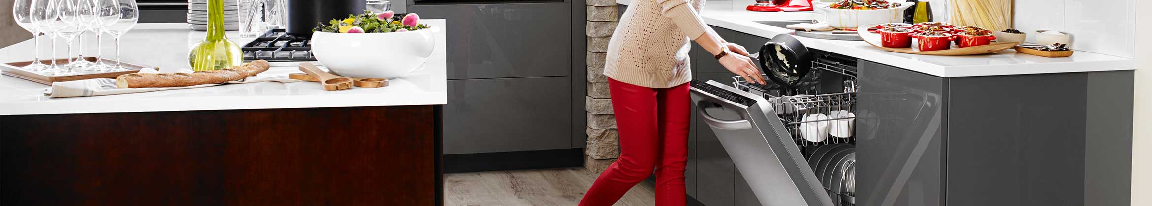 Someone is removing a pot from an opened KitchenAid dishwasher. On the counter in front of them are small crock pots and various other kitchen items. On the island behind them are a salad bowl filled with salad and other kitchen and food items.