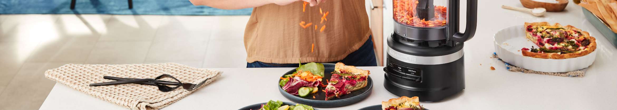 Someone garnishes a salad with shredded carrots from a KitchenAid Food Chopper. On the plate with the salad is a slice of quiche. On the counter is the remaining quiche pie, a folded dish cloth, and a bowl of salt. 