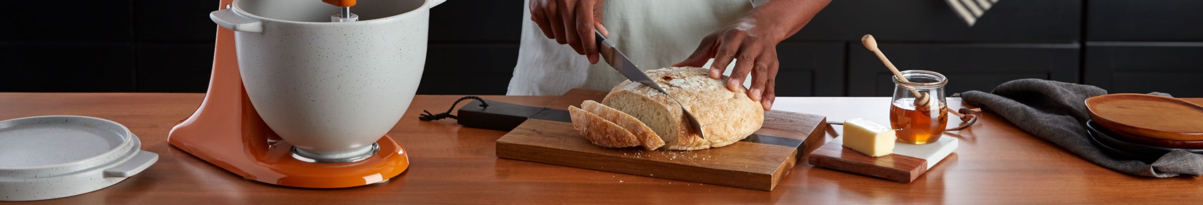 Someone slices bread on a cutting board. A KitchenAid Stand Mixer is on the counter along with a small cutting board with butter and honey and a pie on a dish cloth.