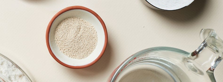 A close-up image displays essential baking ingredients showcasing a measuring cup of water alongside dishes of flour and dry yeast.