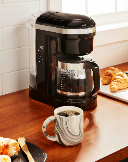 A black KitchenAid coffee maker with a cup of coffee and croissants nearby.