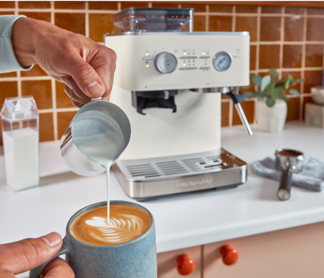 Une personne versant du lait chauffé à la vapeur dans son cappuccino, avec une machine à espresso KitchenAid® en arrière-plan.