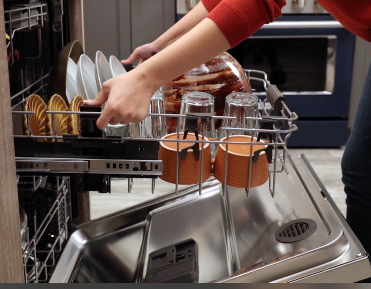 A person grips the sides of a rack that's pulled out from a KitchenAid Dishwasher. On the rack are glasses, plates, ramekins and large bowls