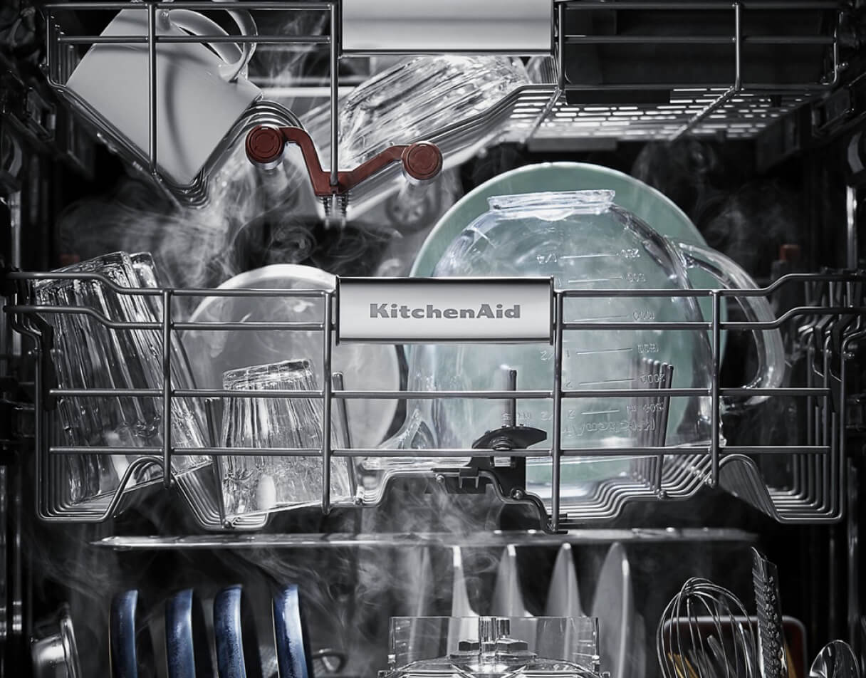 The inside of a KitchenAid Dishwasher. The front of a rack has a plaque that reads, "KitchenAid". The items on the racks include glassware, large bowls and other items