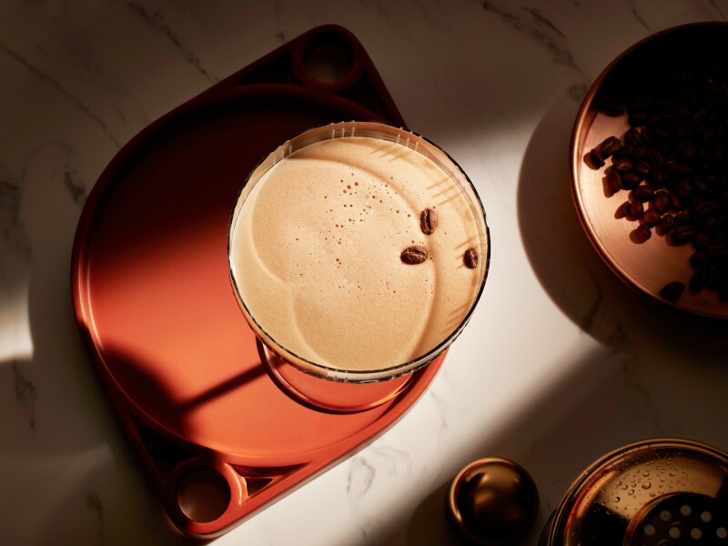 : Bird's eye view of a espresso martini on a brown coaster. Next to it is a dish of coffee grounds. 