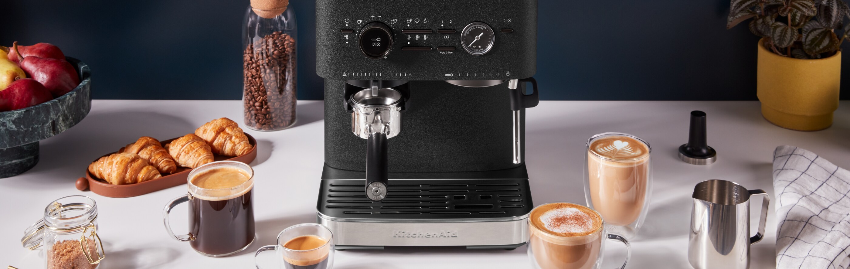 A KitchenAid espresso maker is seen surrounded by a variety of espresso beverages alongside tempting pastries and fruit.