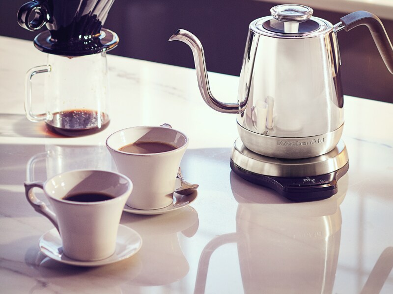 Une bouilloire électrique à bec en col de cygne KitchenAid® en argent sur un comptoir blanc avec un récipient pour le café et deux tasses de café.
