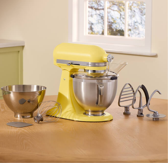 In a kitchen with a window is a Butter KitchenAid Stand Mixer next to a mixing bowl and some attachments. 