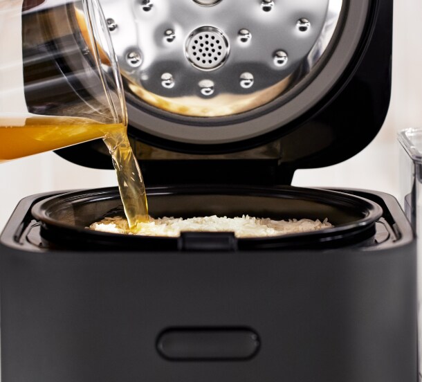 Broth being poured over uncooked white rice in the grain and rice cooker.