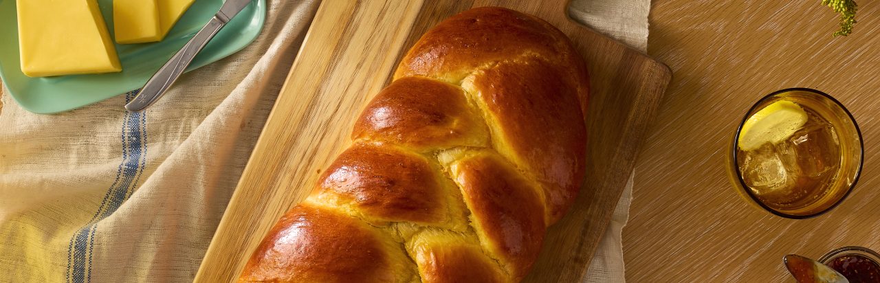 A brioche on a cutting board. Next to it is a butter dish with butter and a knife on it and a glass of water with lemon and ice.  