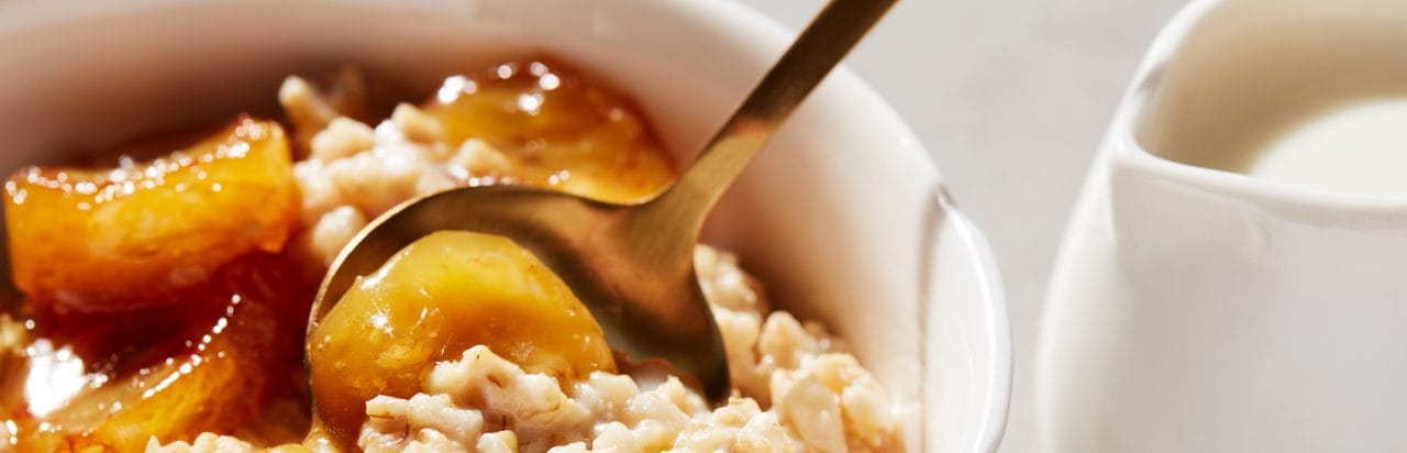 A bowl of creamy oats with caramelized bananas. A spoon is in the bowl and there's a creamer next to the bowl. 