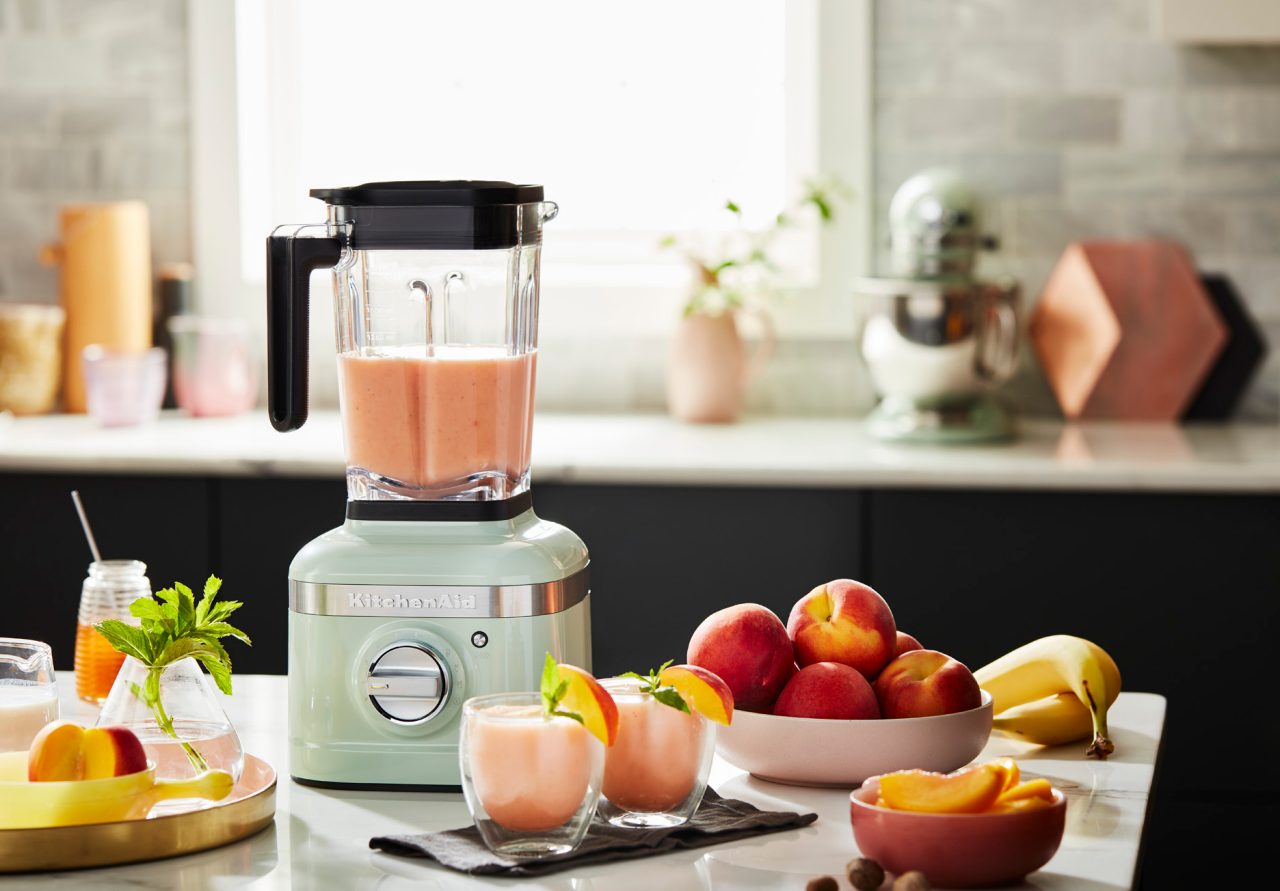 Mélangeur KitchenAid K400 sur le comptoir d’une cuisine à côté de fruits frais et d'ingrédients préparés pour créer une boisson frappée aux pêches et bananes