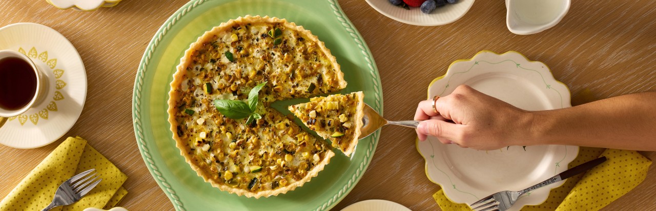 Une personne prend une part de quiche posée sur une table. Sur la table se trouvent également une assiette, un bol avec des fruits, une tasse de thé sur une soucoupe et deux serviettes avec des fourchettes. 