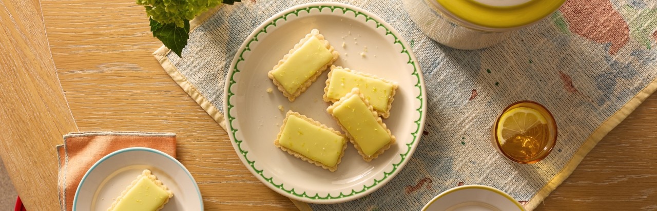 Sur une table se trouve une assiette contenant quatre sablés givrés au yuzu. Sur la table se trouvent aussi un verre d'eau avec un citron et des glaçons, une autre assiette avec un morceau de sablé et différents objets. 