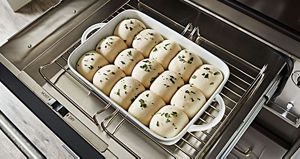 On a rack in a KitchenAid warming drawer is a baking dish with raw dough in the shape of rolls, garnished with herbs