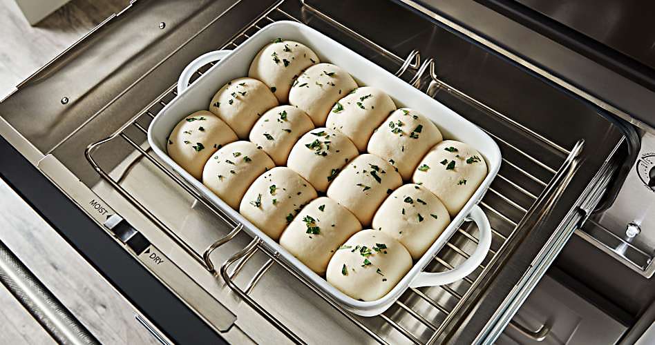 On a rack in a KitchenAid warming drawer is a baking dish with raw dough in the shape of rolls, garnished with herbs