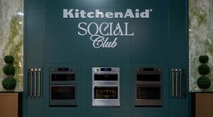 A wall with "KitchenAid Social Club" and three wall ovens. Next to the wall ovens are samples of finishes.