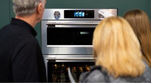 A woman demonstrates how to use a KitchenAid wall oven to two people by turning a knob. A tray of baked goods cooks in the bottom oven. 