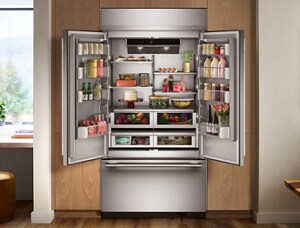 A woman cooks at a KitchenAid oven. In the kitchen is a KitchenAid dishwasher and KitchenAid refrigerator. On the island is a plate of chicken, a salad, and a bowl with berries.