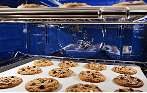 The blue interior of a wall oven, with two racks of chocolate chip cookies baking.