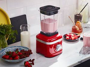 Red K150 Blender with a fresh smoothie, beside a plate of cut strawberries and jar of honey.