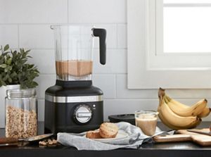 Black K400 Blender with home-made nut butter in it, beside a jar of nuts and a bunch of bananas.